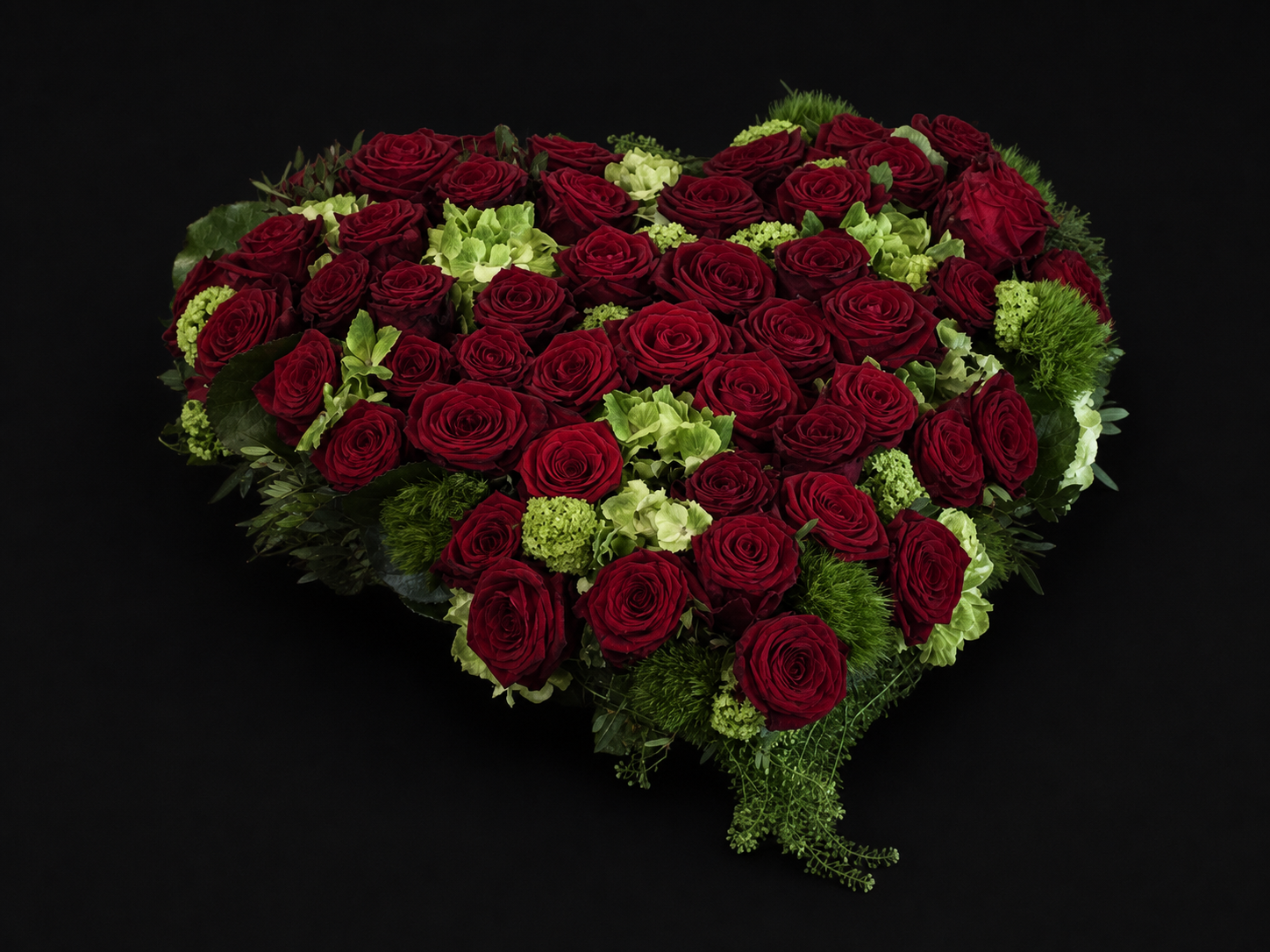 Herzförmiger Trauerschmuck aus dunkelroten Rosen und natürlichem Grün vor schwarzem Hintergrund